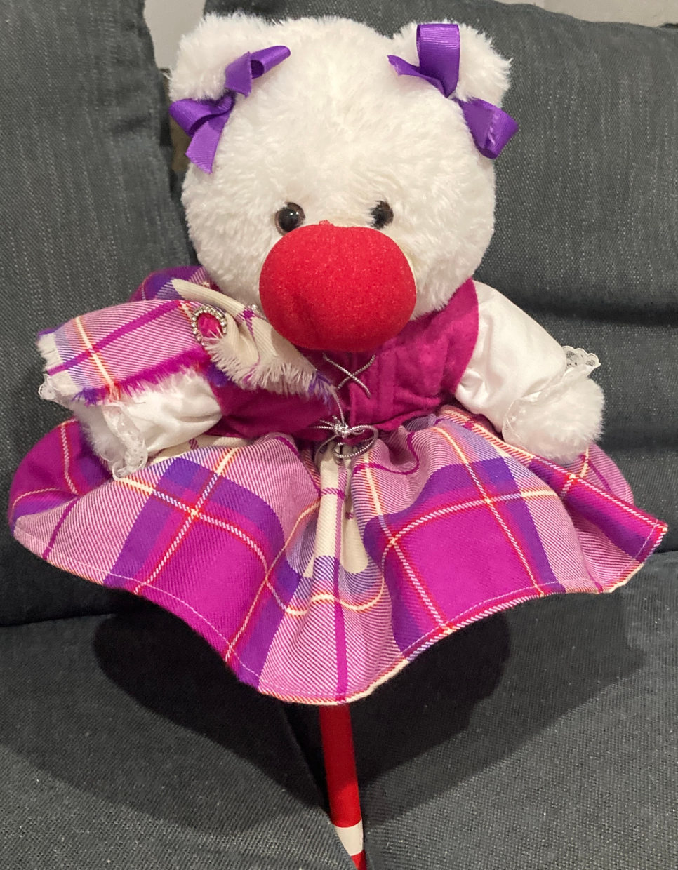 Teddy bear wearing red clown nose and purple/pink flannel dress. Sitting on red plastic stick.