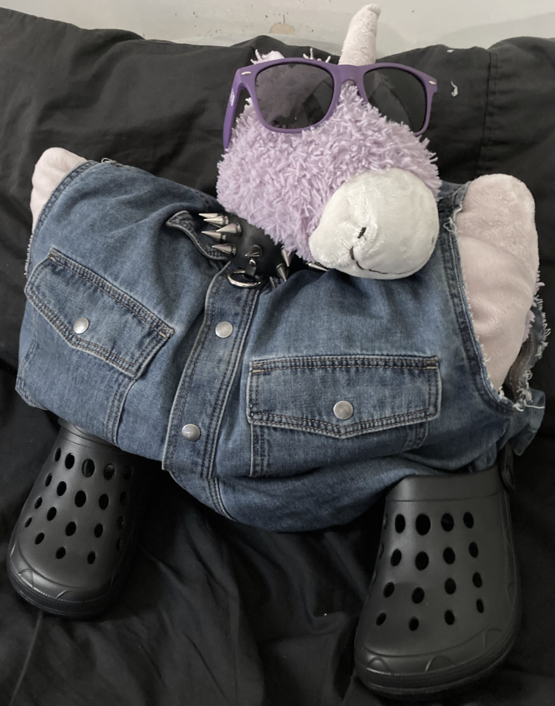Purple unicorn plushy. Denim. black crocs, choker. Purple sunnies. Resting on black bed.