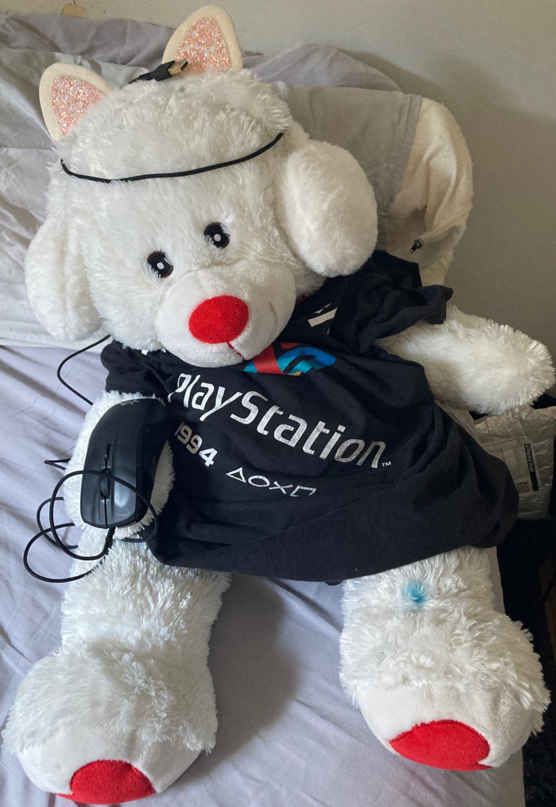 White teddybear wearing black playstation t-shirt, white cat earmuffs, holding computer mouse.