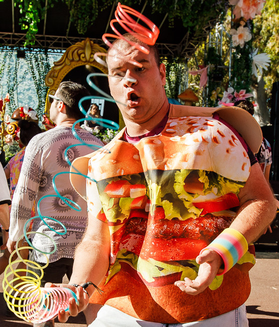 Slinkyfam playing with slinky, shocked face. Burger outfit, denim pants. Daytime, infront of stage at rave.